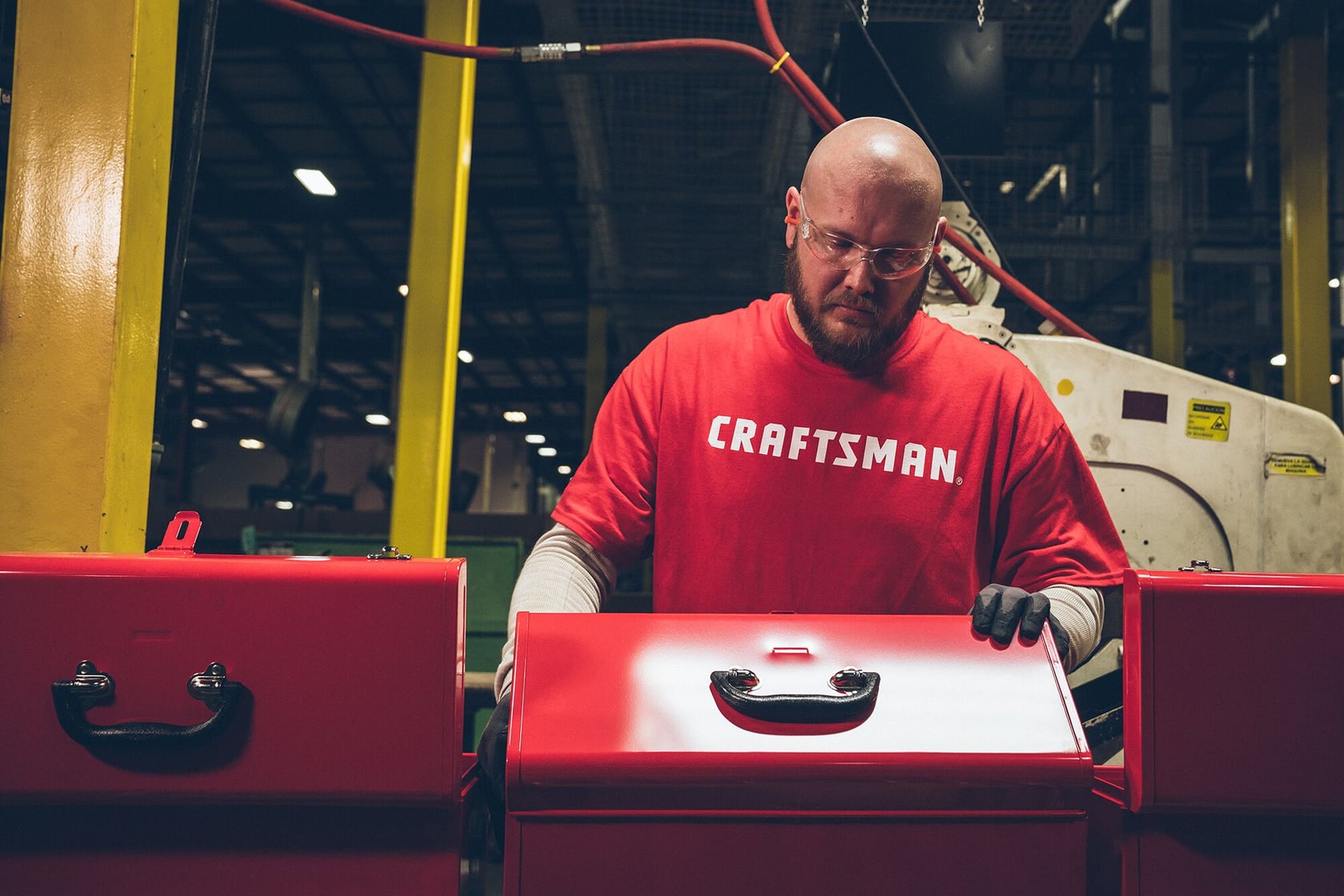 Photo of worker in red CRAFTSMAN® shirt assembling red toolboxes, industrial setting, SKU not visible.