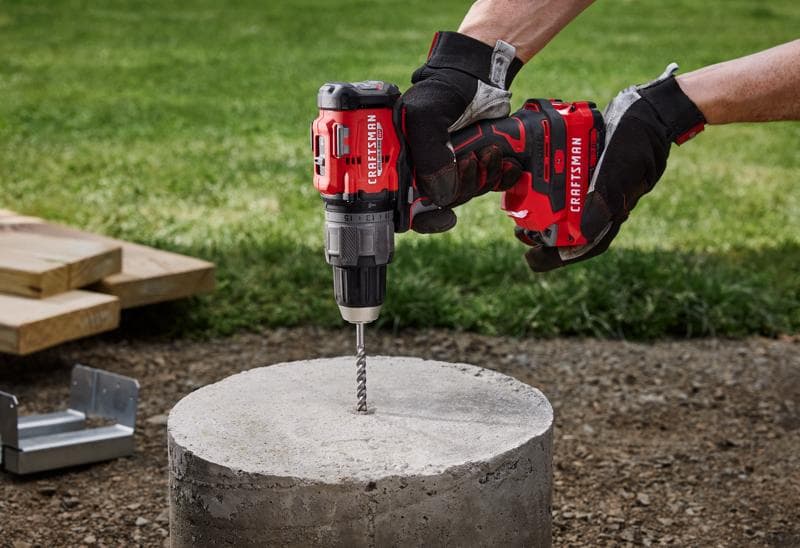 Photo of CRAFTSMAN cordless hammer drill CMCD726B drilling into concrete outdoors, held by gloved hands.