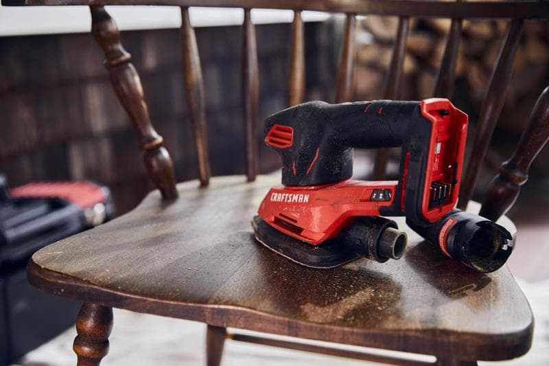 Craftsman cordless sander CMCW221B resting on a wooden chair, red and black, with dust port attachment.