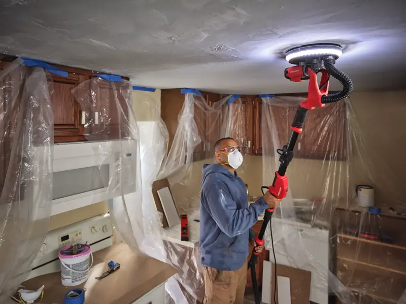 Person uses CRAFTSMAN pole sander on ceiling in kitchen with cabinets covered in plastic sheets.