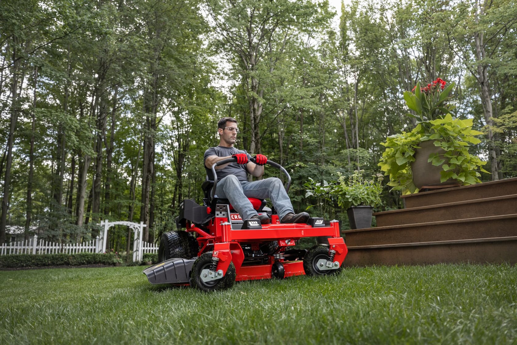 Photo of a person operating a red CRAFTSMAN zero-turn lawn mower, SKU CMGN231702, on grass beside garden steps.