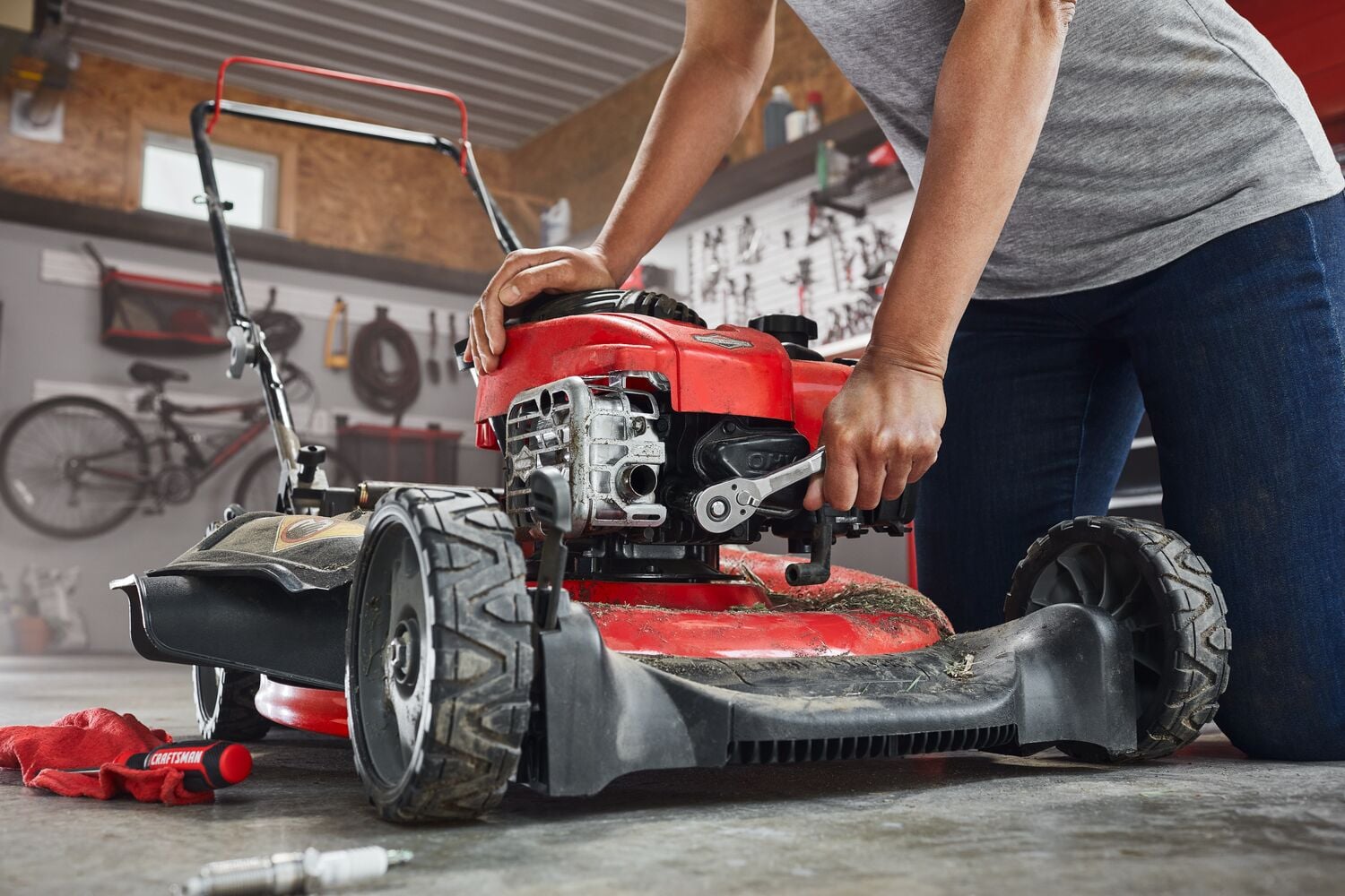 A person is performing maintenance on a red lawn mower in a garage, using a wrench to work on the engine. Various tools and a red cloth are visible on the floor.