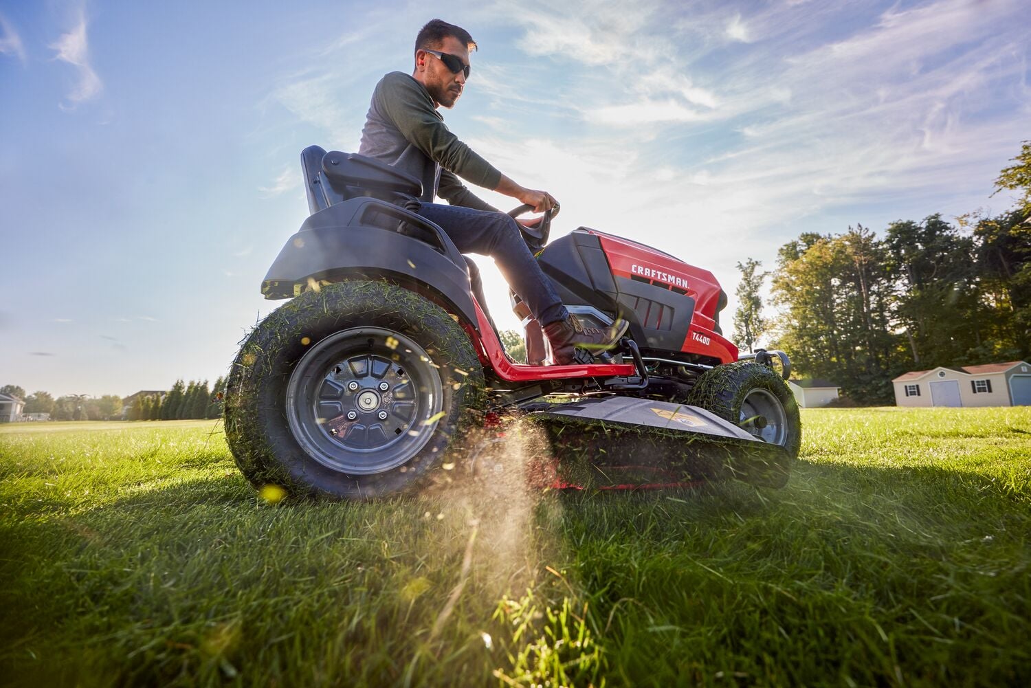 A person operating a red Craftsman riding lawn mower on a grassy field under a blue sky, with grass clippings being discharged from the side of the mower.