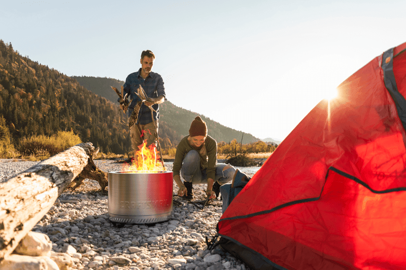 Two people are setting up a wood fire in a portable metal fire pit next to a bright red tent on a rocky surface, with logs and mountains in the background under a clear sky.