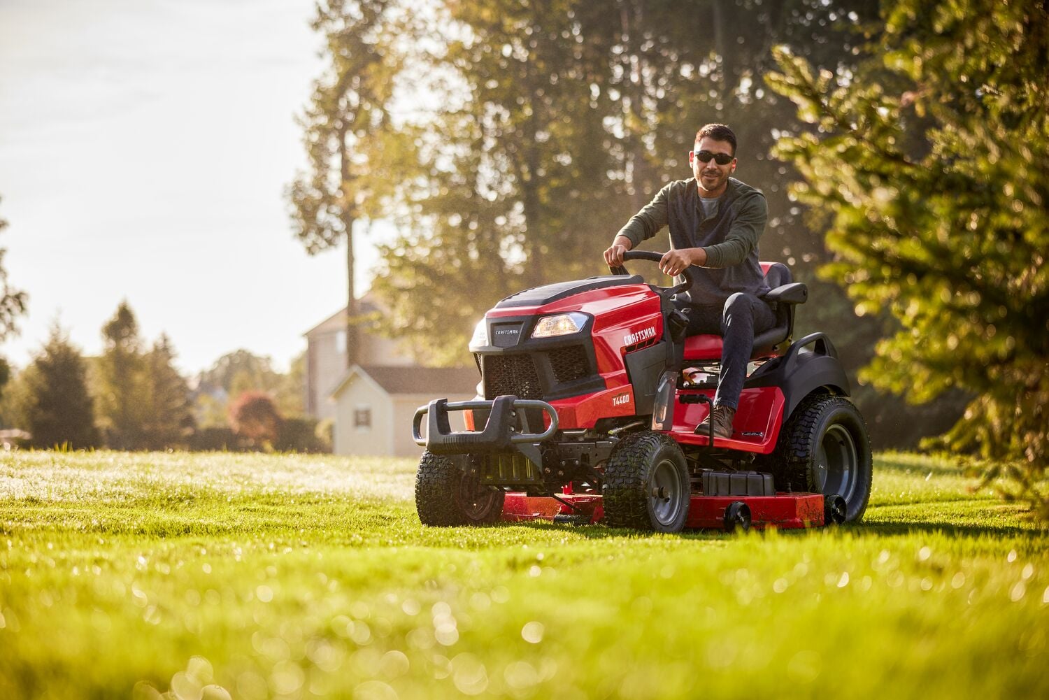 A person is riding a red Craftsman riding lawn mower on a sunny lawn with trees and a house in the background.