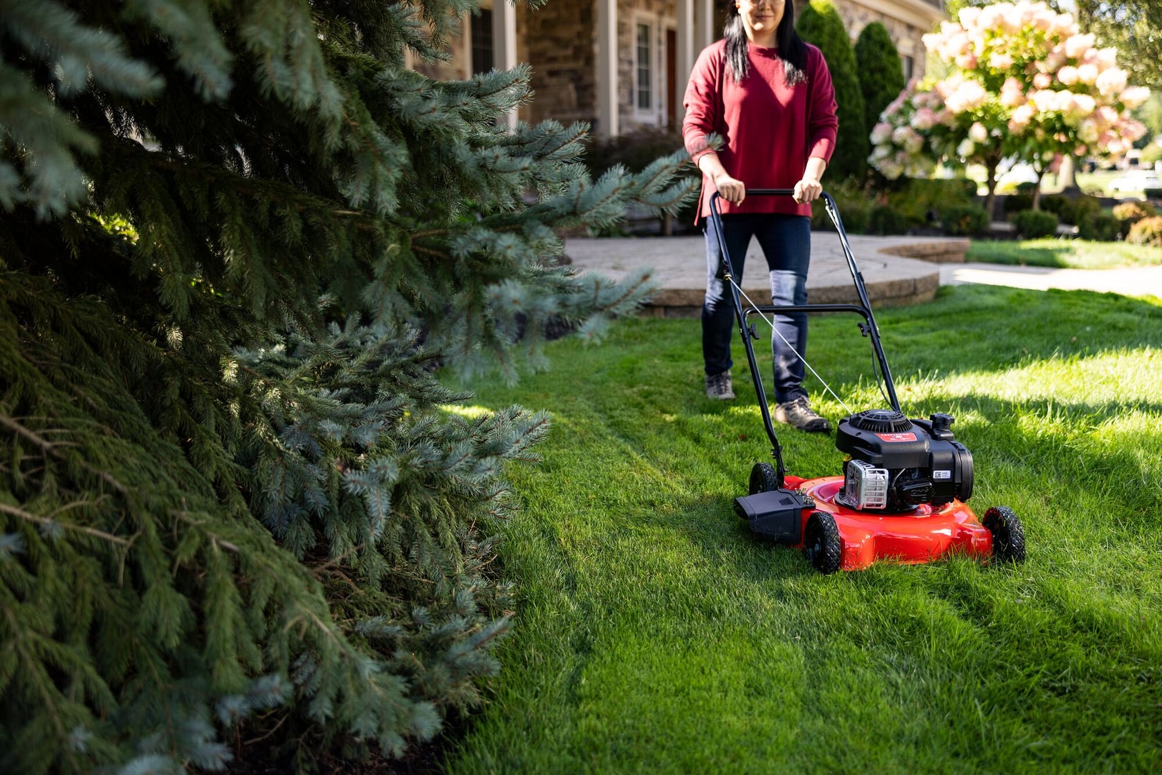 A person using a red and black gas-powered lawn mower to cut grass in a residential yard, with a house and trees in the background.
