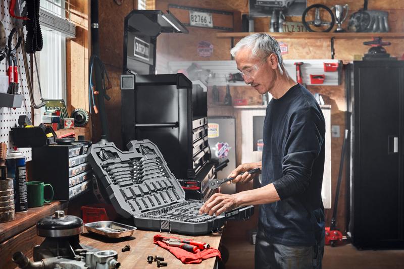 A person is working in a garage or workshop, using a wrench and standing at a workbench with an open tool case containing various sockets and tools. The workspace is filled with automotive parts, toolboxes, and equipment.
