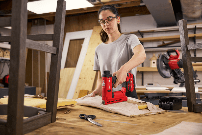 Woman using CRAFTSMAN V20* stapler to attach fabric to barstool seat in a woodshop setting.