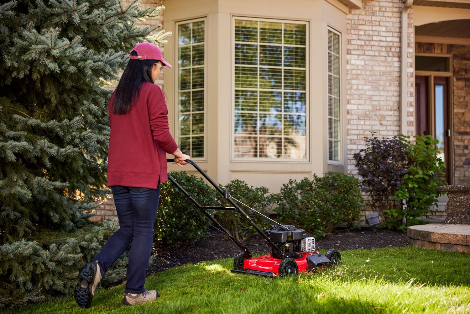 A person wearing a pink cap and red sweater is pushing a red lawn mower on a green lawn near a house with large windows and landscaping including bushes and a tree.
