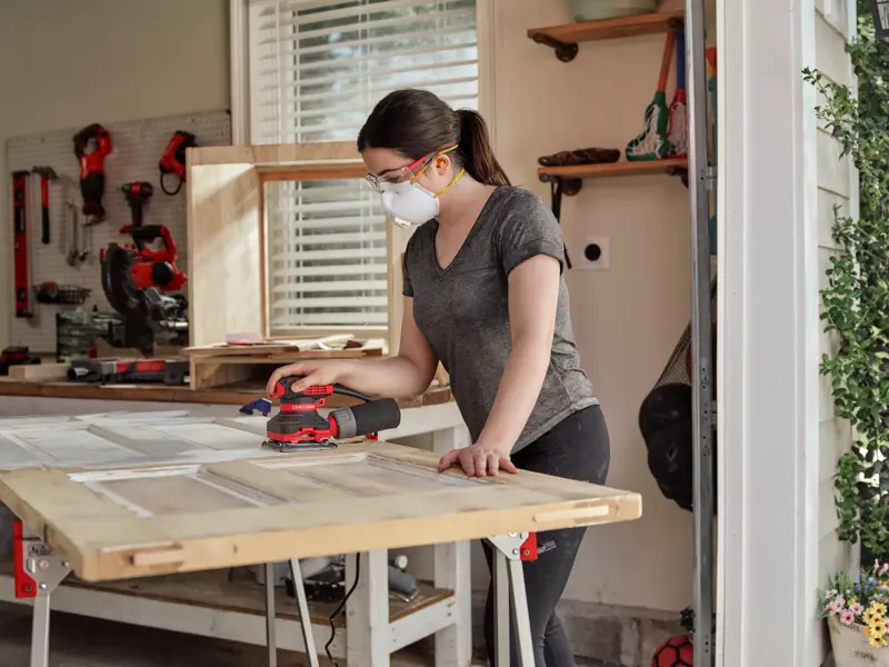 Person uses a CRAFTSMAN sander on wood, wearing a mask and goggles, in a workshop with tools visible in the background.