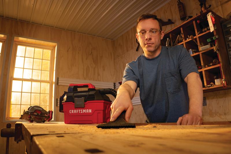 Photo of a person using a CRAFTSMAN CMCV002B wet/dry vacuum on a workbench in a woodshop, vacuum cleaning sawdust.