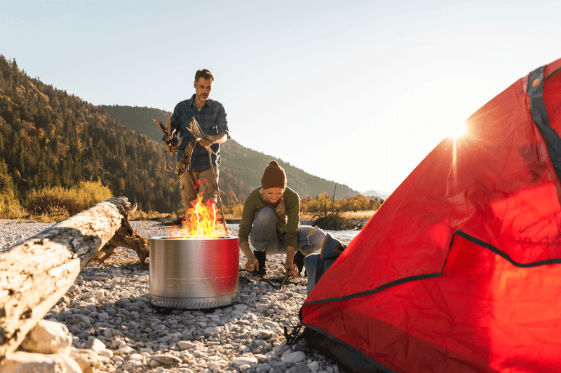 Two people are setting up a wood fire in a portable metal fire pit next to a bright red tent on a rocky surface, with logs and mountains in the background under a clear sky.