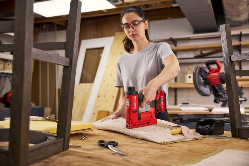 Woman using CRAFTSMAN V20* stapler to attach fabric to barstool seat in a woodshop setting.