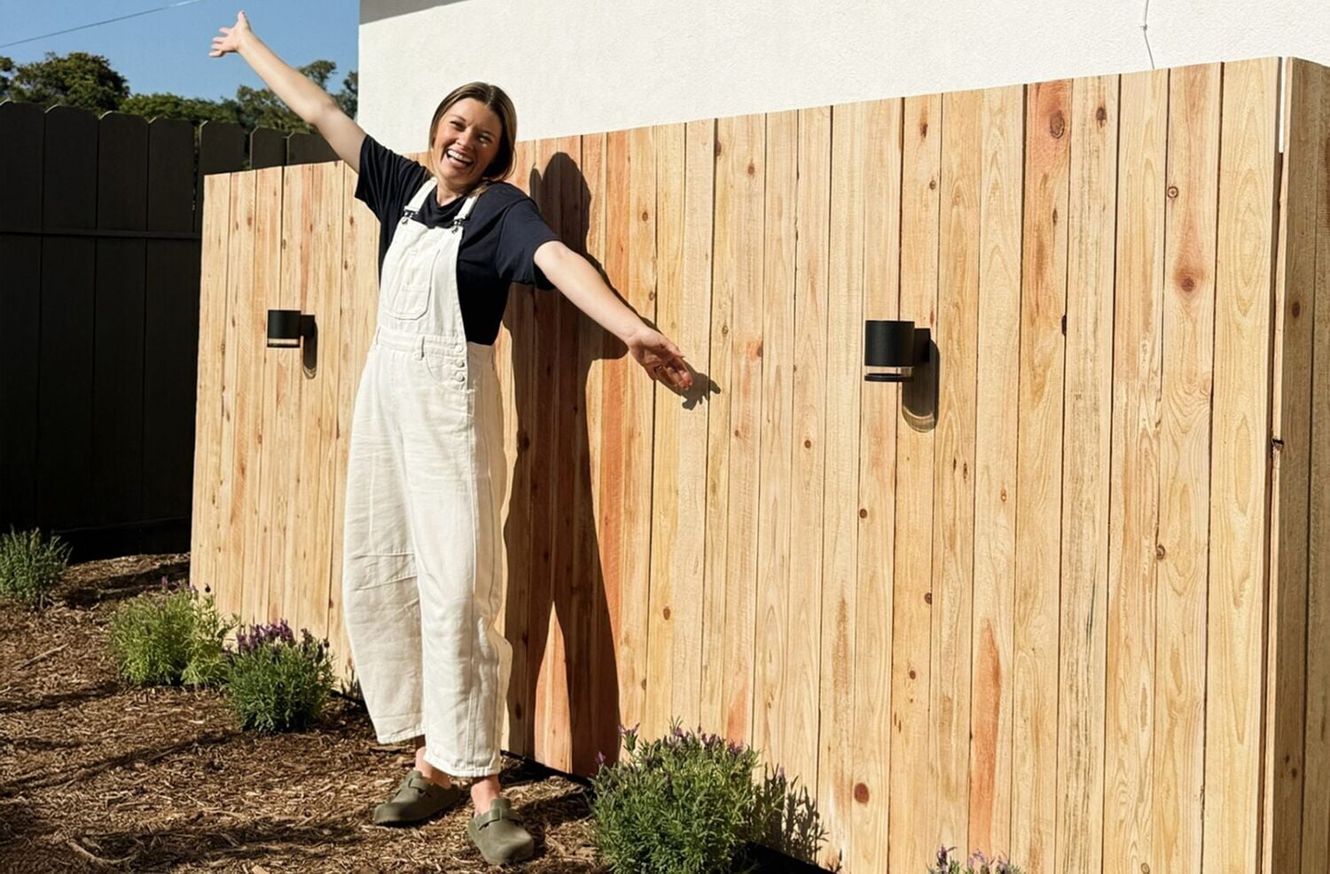 Person wearing light-colored overalls and dark shirt standing with arms outstretched in front of a wooden fence with mounted black outdoor lights. There are small plants in the soil near the fence.