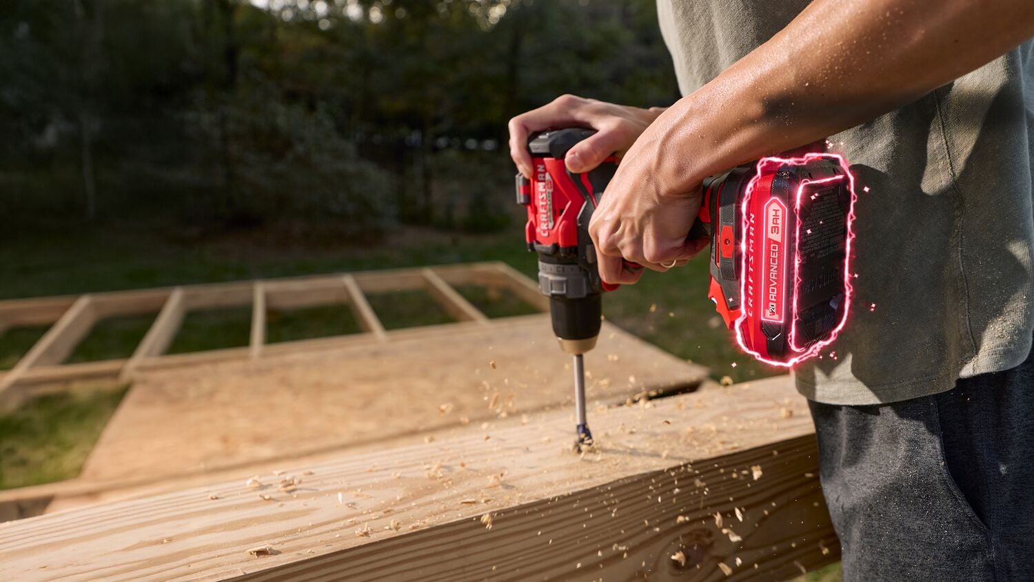 A person using a red CRAFTSMAN cordless drill to drill into a wooden plank outdoors. Wood shavings are flying as the drill bit penetrates the wood. The drill’s battery is highlighted with a glowing effect.