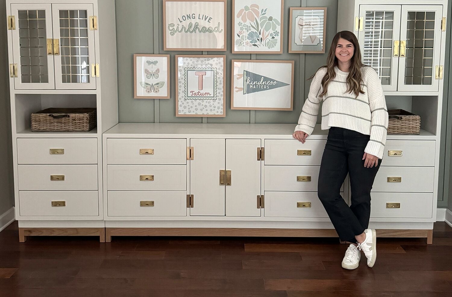 A person wearing a white sweater and black pants is leaning against a large cream-colored cabinet with brass handles. The cabinet features drawers and glass doors, and there are wicker baskets placed inside open shelves. Above the cabinet, there are framed decorative wall art pieces with pastel designs and phrases. The floor is dark wood.