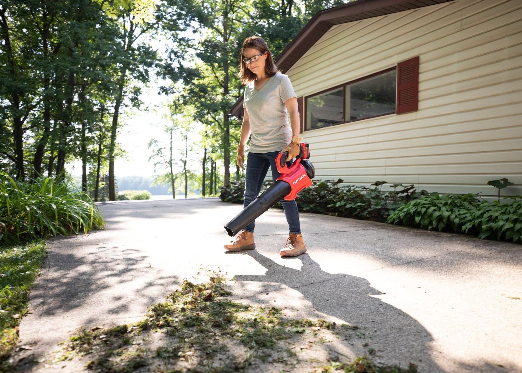 Photo of a person using a CRAFTSMAN cordless leaf blower, SKU CMCBL730P1, on a driveway next to a house.