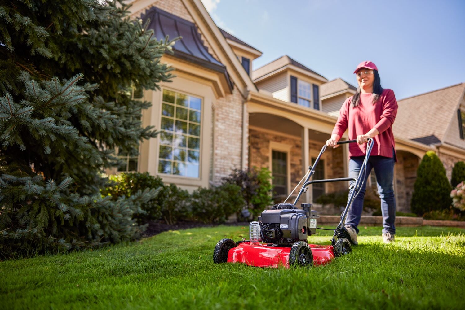 Person using a red lawn mower to cut grass in front of a suburban house on a sunny day.