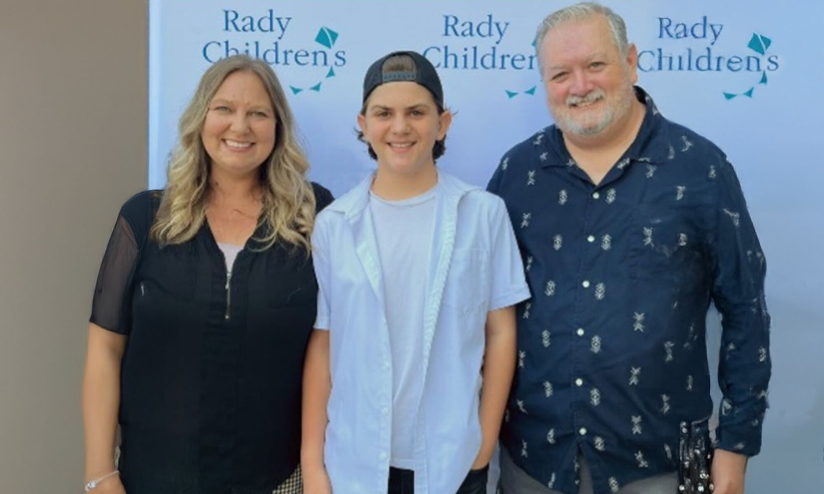 Three people with blurred faces standing in front of a backdrop that reads 'Rady Children's.' The person on the left is wearing a black shirt, the person in the middle is wearing a light shirt and a dark cap, and the person on the right is wearing a dark patterned shirt.