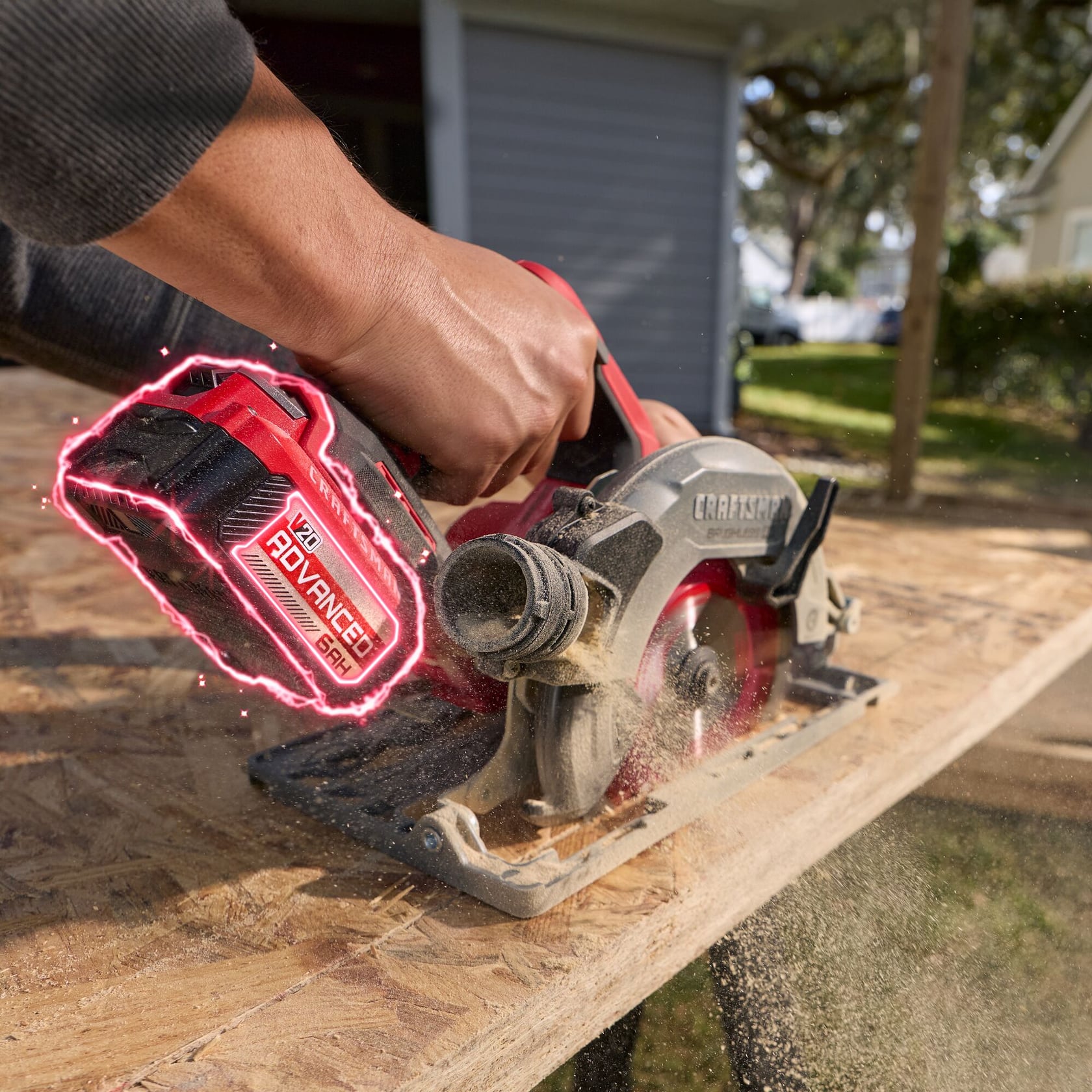 A close-up of a person's hand operating a CRAFTSMAN cordless circular saw with a red V20 ADVANCED 6.0Ah battery, cutting through a sheet of plywood outdoors. Sawdust is visible flying from the wood.