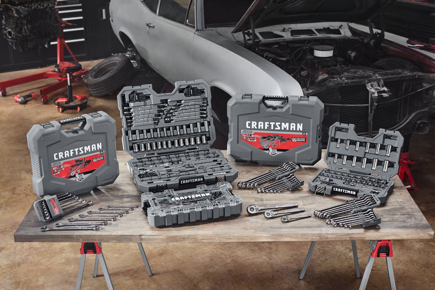 A table in a garage displaying several open and closed Craftsman tool cases, filled with various hand tools including wrenches, sockets, and ratchets. An old car with its hood open is in the background, along with automotive equipment and spare tires.