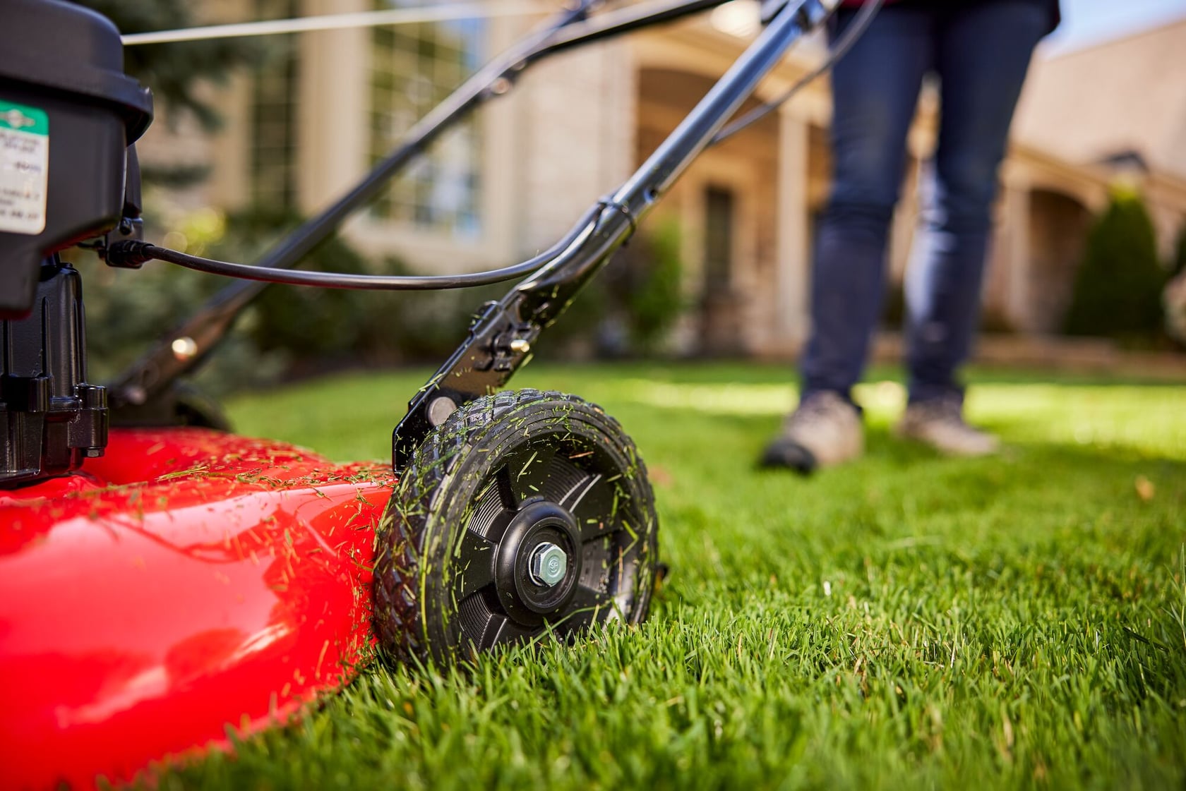 Close-up view of a red lawn mower cutting green grass, with a person standing behind and a house visible in the background.