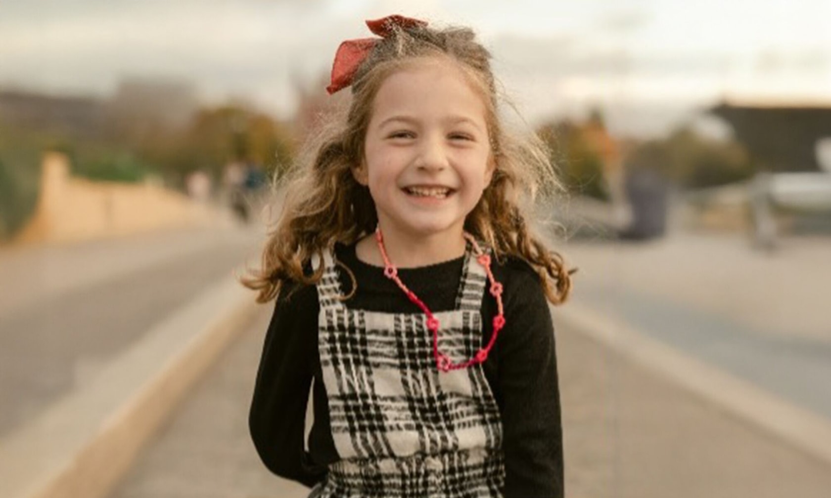 A child with long curly hair, wearing a plaid dress over a black shirt, a red necklace, and a red bow in their hair, standing outdoors on a paved path. The face of the child is blurred for privacy.