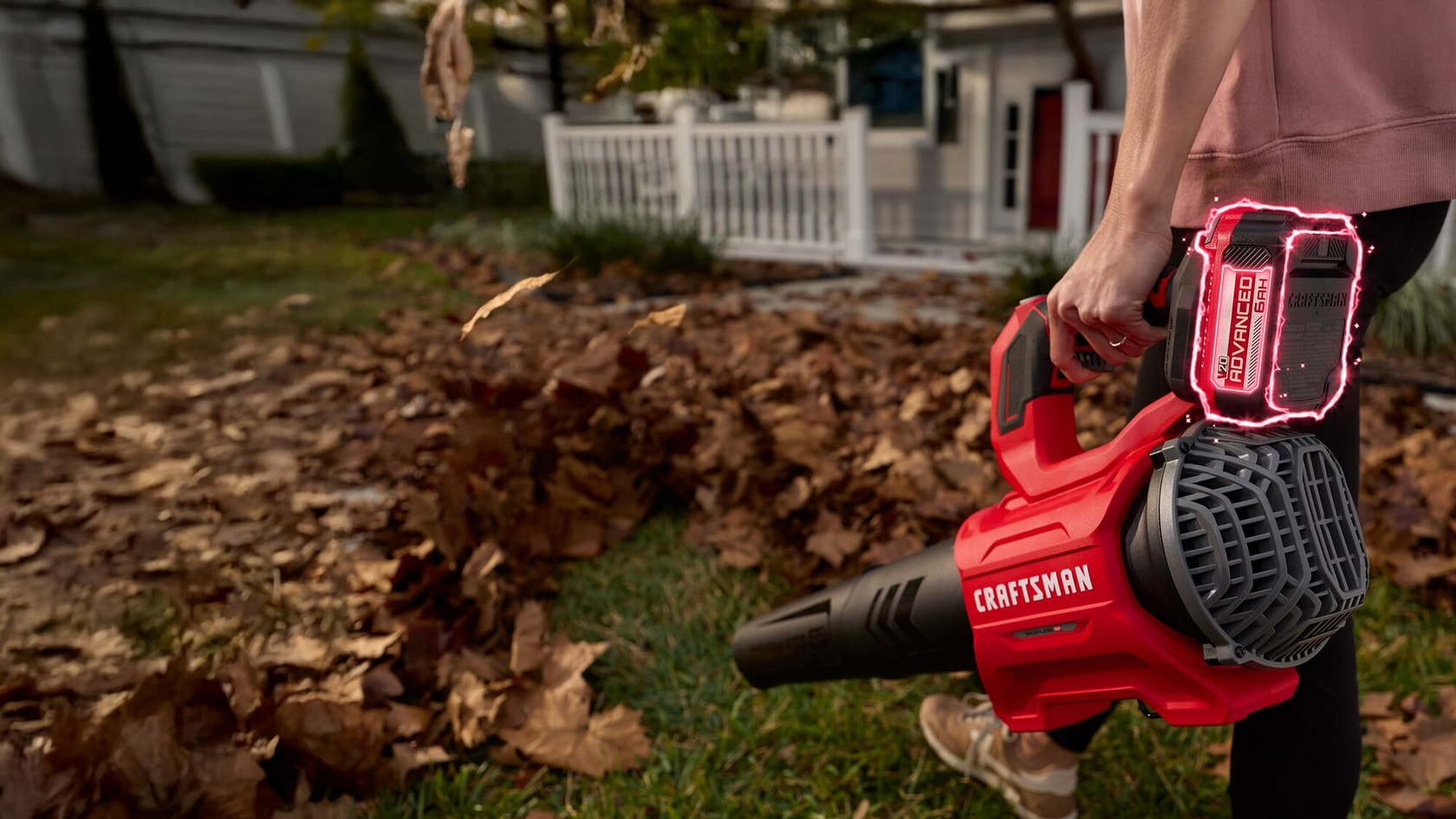 A person using a red CRAFTSMAN cordless leaf blower, model CMCB2103-CK, to clear leaves from a lawn in front of a house with a white fence.
