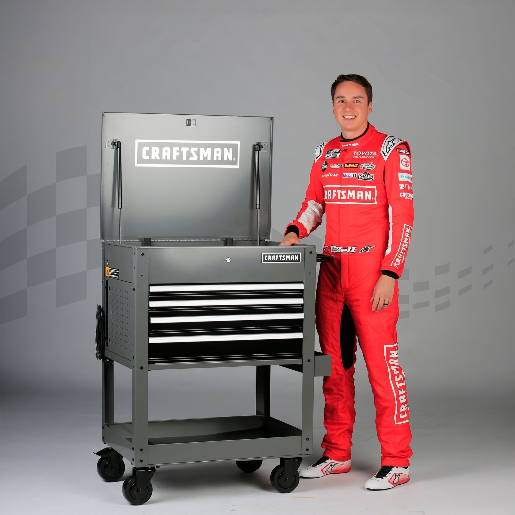 A person in a red racing jumpsuit stands next to a CRAFTSMAN tool chest on wheels with the lid open. The tool chest has multiple drawers and a prominent CRAFTSMAN logo.
