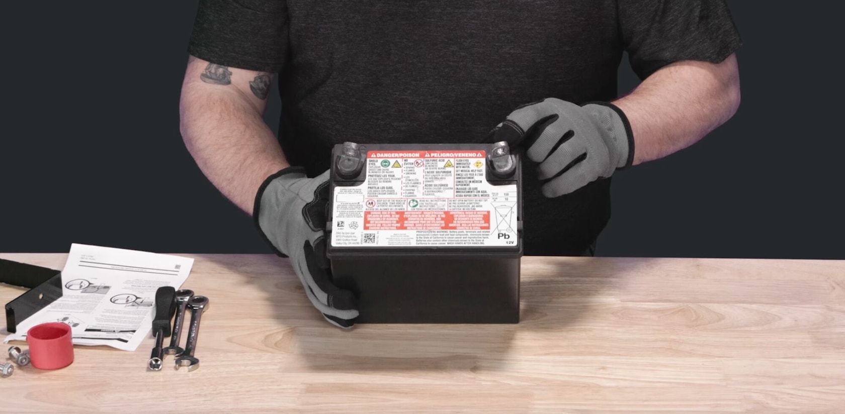 Craftsman lawn mower battery held by gloved hands, manuals and tools on wooden table, front angle.