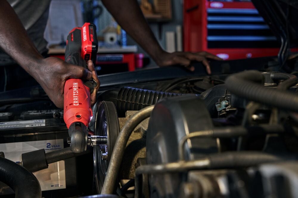 Photo of a person using a red Craftsman cordless ratchet tool, SKU CMCF935, while working on a car engine.