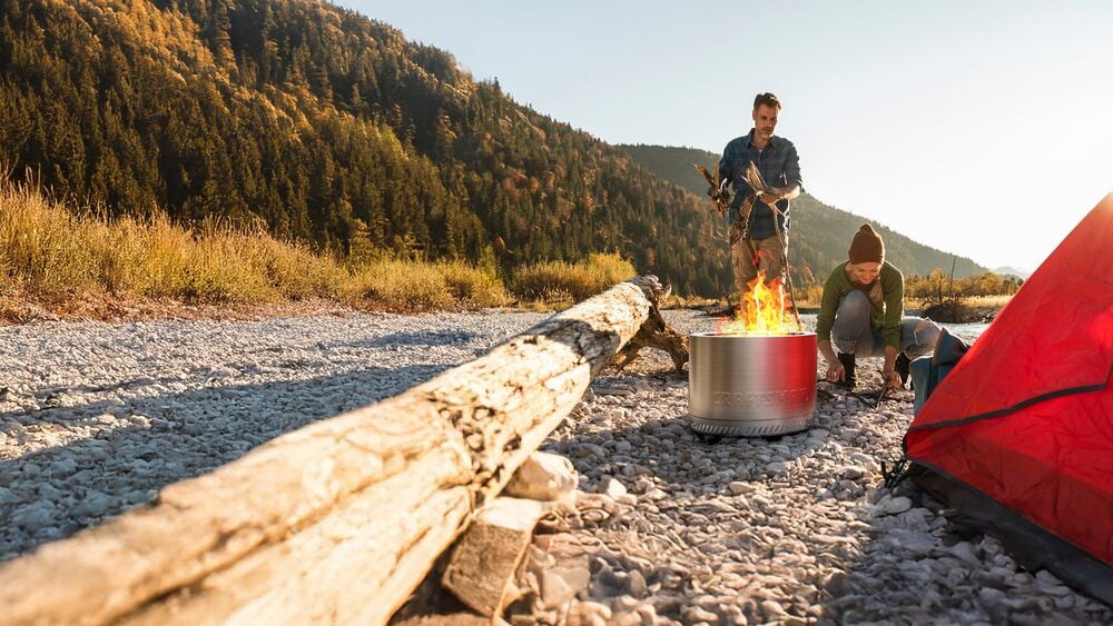 Photo of two people by a CRAFTSMAN stainless steel fire pit, SKU visible, next to a red tent in a mountain setting.