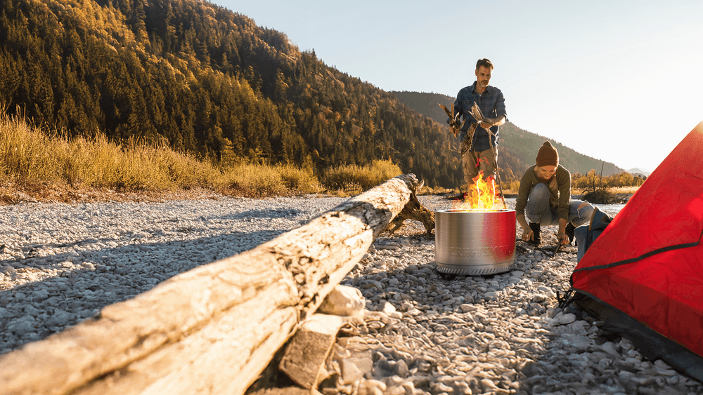 Outdoor camping scene with a metal fire pit burning on rocky ground, surrounded by logs, a red tent, and scenic forested hills.