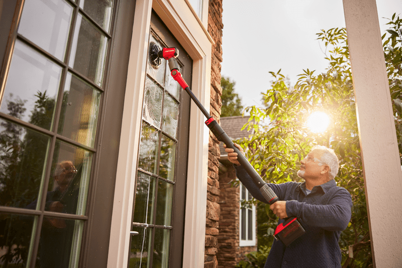 Person using a cordless electric cleaning tool with an extension pole to clean exterior windows of a house, with sunlight visible in the background.